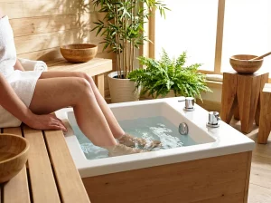 Person sitzt in einem Spa-Bereich auf einer Holzbank und nimmt ein Fußbad in einem quadratischen, weiß emaillierten Kneipp-Fußbecken mit Holzverkleidung.